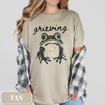 Tan Bella Canvas 3001 T-shirt with a large frog graphic that says "Grieving". A way for people struggling with loss to convey their sorrow - makes a perfect gift for a friend or loved one struggling with grief.