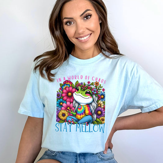 Chambray comfort colors t-shirt with an adorable hippie frog with big eyes, wearing a tie dye shirt and blue jeans, surrounding by a flower garden, and the saying In A World of Chaos, Stay Mellow. 